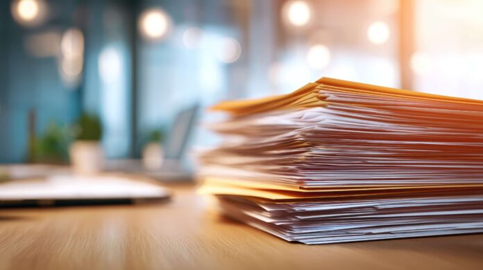 A stack of paper documents and folders sits on a wooden desk in a bright, modern office environment